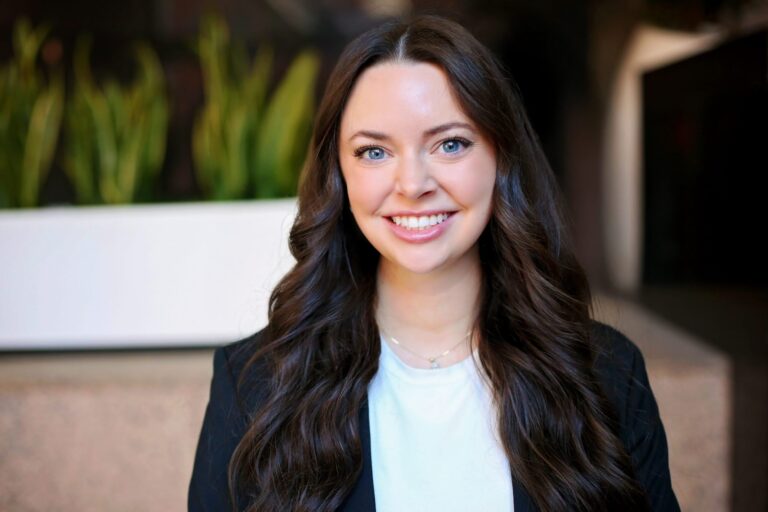 Young woman with long, wavy dark brown hair and blue eyes, smiling. She is wearing a white top and a black blazer, with a delicate necklace. The background is softly blurred with green plants and neutral tones.