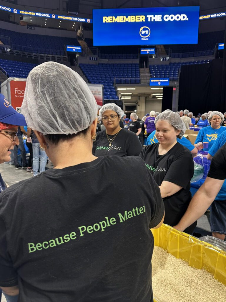 A group of people wearing hairnets and black shirts with "Because People Matter" and "SWMW LAW" printed on them are gathered around a large container filled with grains, likely participating in a food packing or volunteer event inside a large indoor arena. In the background, a large blue screen displays the message "REMEMBER THE GOOD." with a 9/11 logo. Other volunteers in blue shirts are also visible.