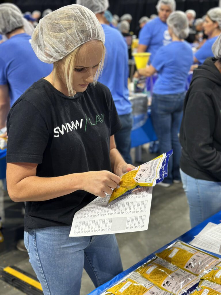 A woman wearing a black "SWIMMI/LAND" t-shirt, light blue jeans, and a hairnet is examining a yellow and blue packaged item while holding a checklist or form. She is standing at a table covered with similar packaged items, and other people in blue shirts and hairnets are visible in the background, suggesting a group activity or volunteer event.