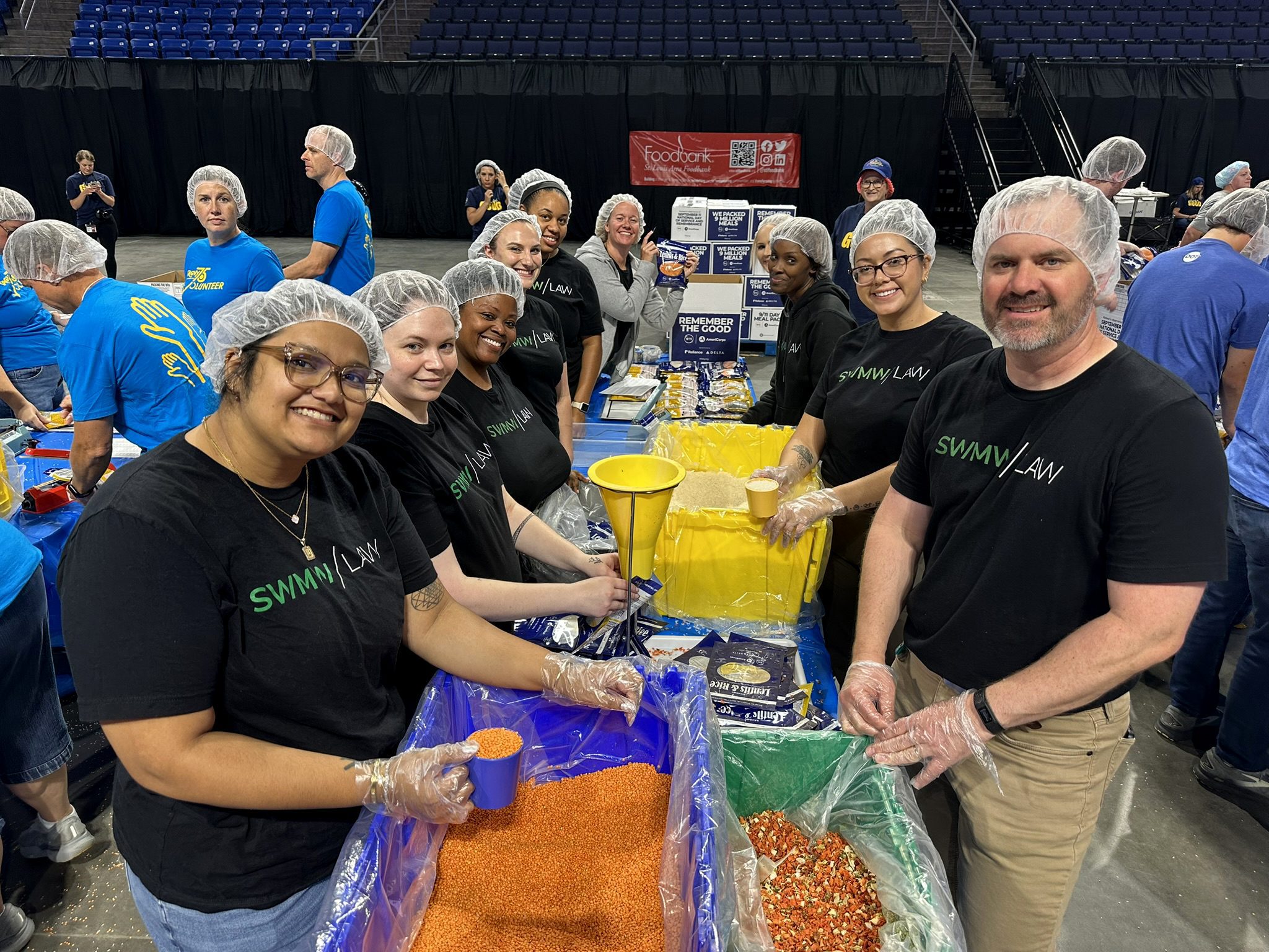 A group of volunteers wearing black "SWMW LAW" t-shirts and hairnets are standing around tables with large bins of food ingredients, including red lentils, rice, and a mixed vegetable blend. They are scooping the ingredients into small blue cups, likely as part of a food packing event. Other volunteers in blue shirts are visible in the background. The setting appears to be an indoor arena or large hall.