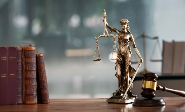 A bronze statue of Lady Justice holding balanced scales and a sword, positioned on a wooden surface. To the left, there are several old, leather-bound books standing upright. To the right, a wooden judge's gavel rests on its sound block. The background is blurred, suggesting an office or courtroom setting.