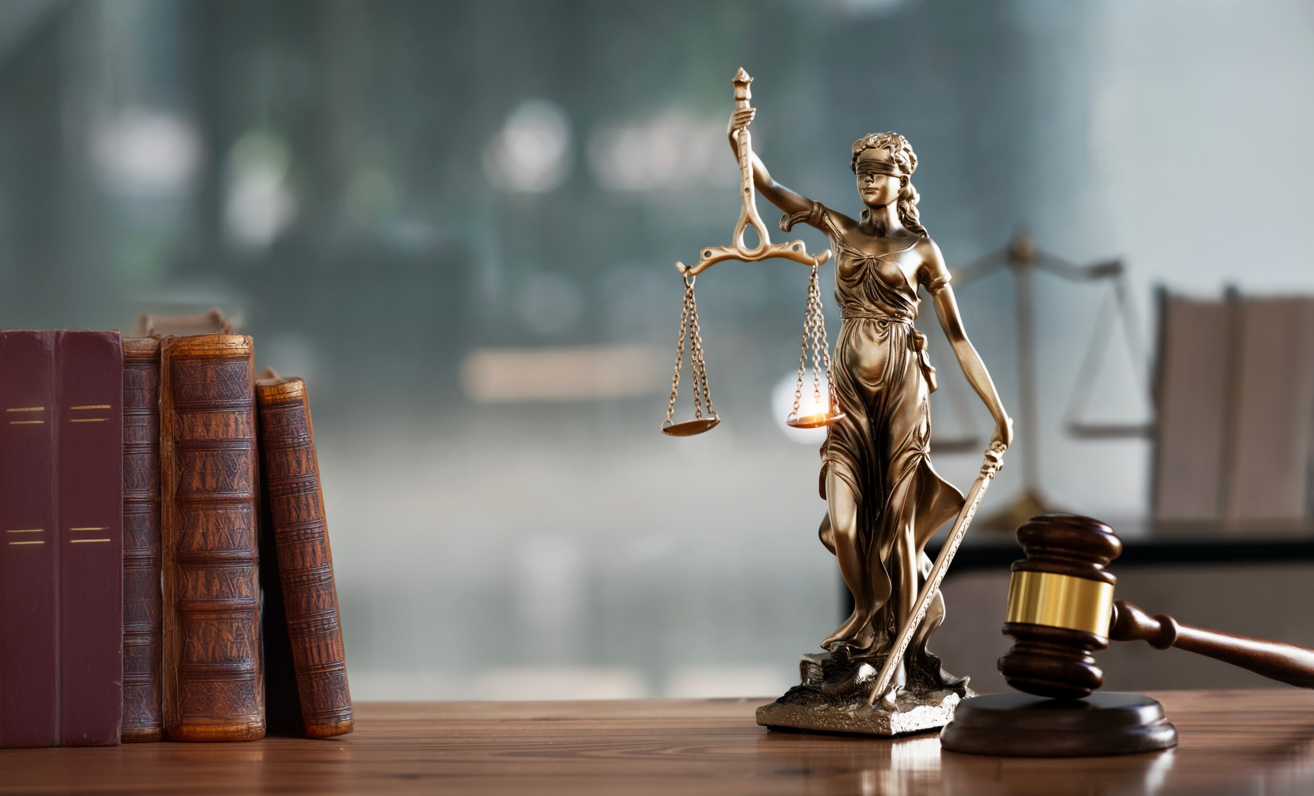 A bronze statue of Lady Justice holding balanced scales and a sword, positioned on a wooden surface. To the left, there are several old, leather-bound books standing upright. To the right, a wooden judge's gavel rests on its sound block. The background is blurred, suggesting an office or courtroom setting.