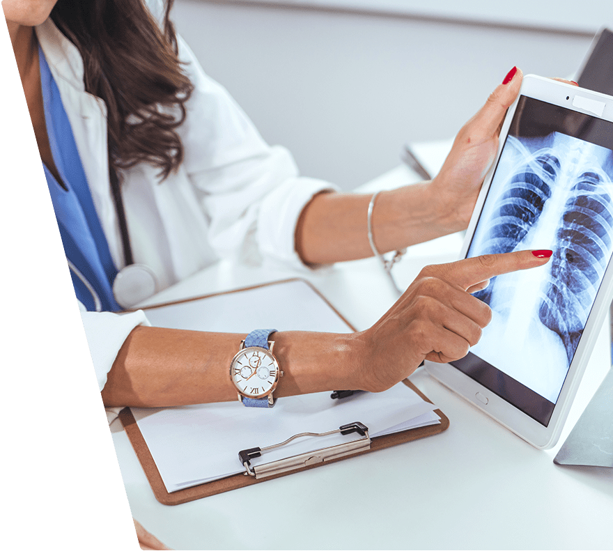 A medical professional wearing a white coat and blue scrubs is pointing at a chest X-ray displayed on a tablet. The X-ray shows the ribcage and lungs. The person has red nail polish and is wearing a wristwatch with a light blue strap. A clipboard with papers is on the table in front of them.