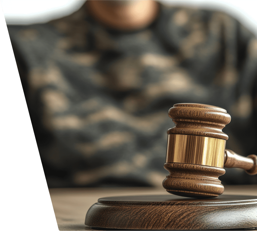 A wooden judge's gavel with a gold band rests on a matching wooden sound block, with a person wearing a camouflage-patterned shirt blurred in the background.