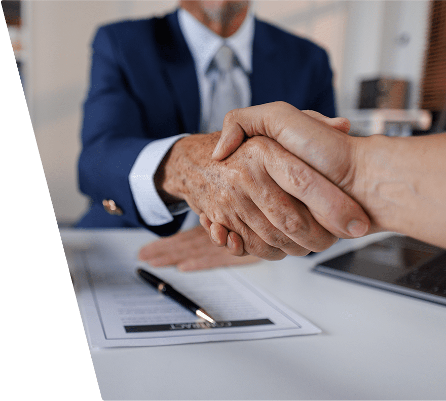 Two people shaking hands over a desk with a contract document and a pen, one person wearing a blue suit and the other with a bare arm.