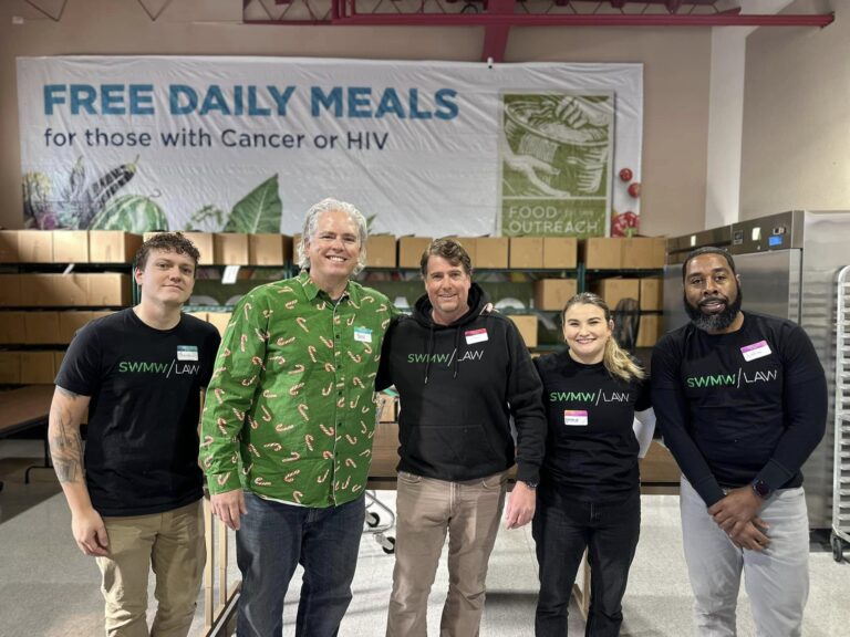 Five people stand side by side indoors in front of a large banner that reads "FREE DAILY MEALS for those with Cancer or HIV." Three individuals wear black shirts with "SWMW | LAW" printed on them, while one person wears a green shirt with a candy cane pattern, and another wears a black hoodie with the same "SWMW | LAW" logo. They are smiling and appear to be part of a volunteer or outreach group. The background includes shelves with boxes and a stainless steel refrigerator.