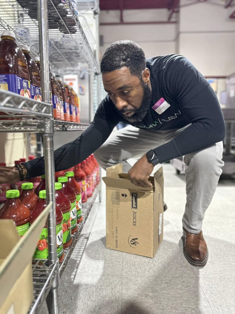 A man with a beard, wearing a black long-sleeve shirt, light gray pants, and brown shoes, is crouching down while stocking or organizing bottles of V8 vegetable juice on metal shelves. He is holding a cardboard box labeled "Campbell's Juices V8" and has a name tag on his shirt. The setting appears to be a store or storage area.