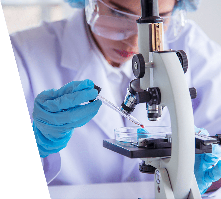 Scientist wearing blue gloves and protective eyewear using a dropper to place a red liquid sample into a petri dish under a microscope.