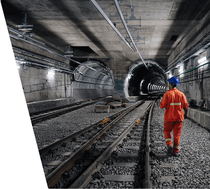 A worker wearing an orange safety uniform and blue helmet walks along railway tracks inside a concrete tunnel under construction. The tunnel has two parallel tracks with gravel between them, and the walls and ceiling are lined with cables and lighting fixtures. The tunnel extends into the distance with lights illuminating the far end.