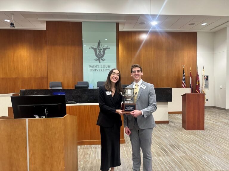 Two people dressed in formal attire stand in a courtroom holding a large silver trophy. The woman on the left wears a black suit, and the man on the right wears a light gray suit with a yellow tie. Behind them is a wooden panel wall with a glass section displaying the Saint Louis University logo and text. There are several flags and a podium to the right, and a computer monitor on a wooden desk to the left. Both individuals have name tags labeled "B-1" and "B-2.