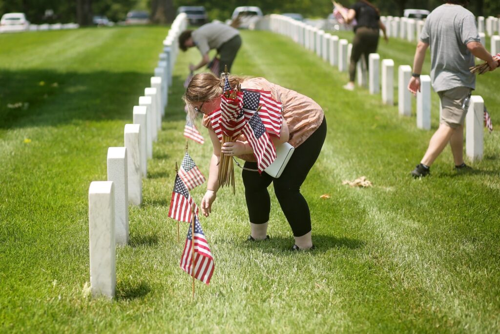 A woman is placing small American flags in front of white gravestones arranged in rows on a grassy lawn. She holds several flags in one arm while bending down to place one. Other people in the background are also placing flags along the rows of gravestones. The scene appears to be a cemetery on a sunny day.