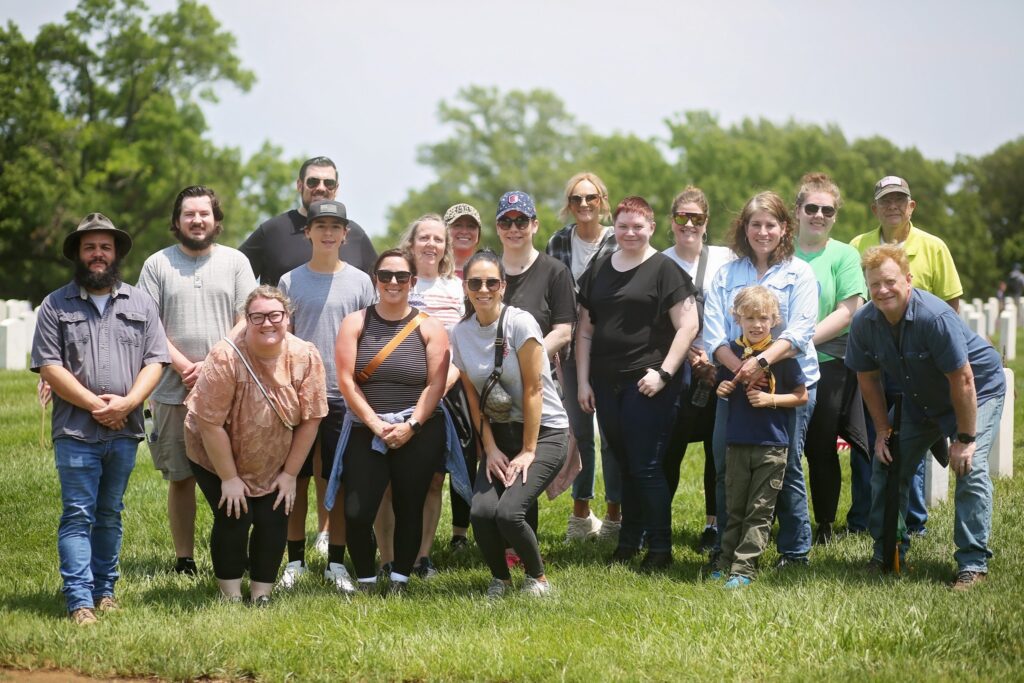 A group of 18 people, including men, women, and a child, posing outdoors on a grassy area with trees and white headstones in the background, suggesting a cemetery setting. Most are smiling, some wearing sunglasses and casual clothing, with a few people crouching in the front row. The weather appears sunny and bright.