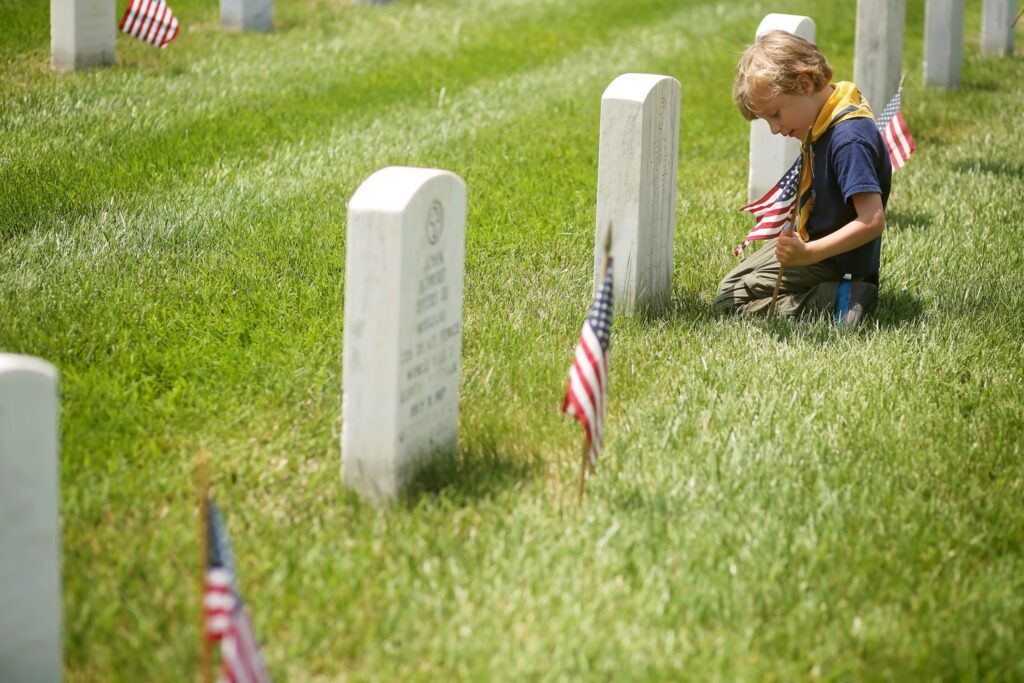 A young boy kneeling on grass in a cemetery, placing an American flag next to a white gravestone. Several other white gravestones and American flags are visible in the background. The boy is wearing a navy blue shirt, khaki pants, and a yellow neckerchief.