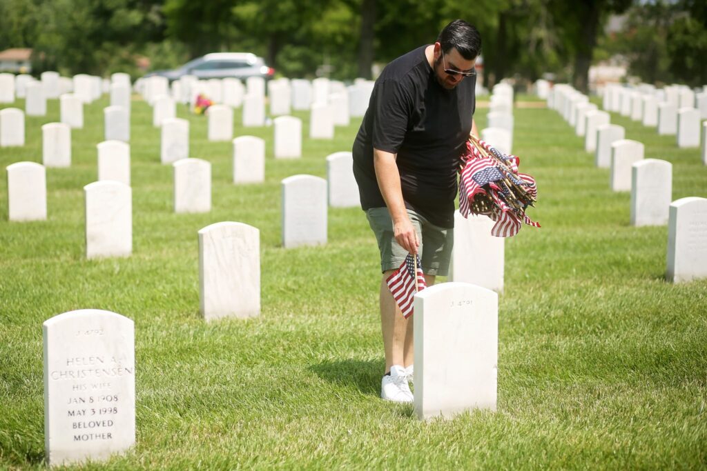 A man wearing a black t-shirt, green shorts, white sneakers, and sunglasses is placing small American flags at a white gravestone in a cemetery. He holds a bundle of similar flags in his left arm. The cemetery has rows of white gravestones on green grass, with trees and a blurred car in the background.