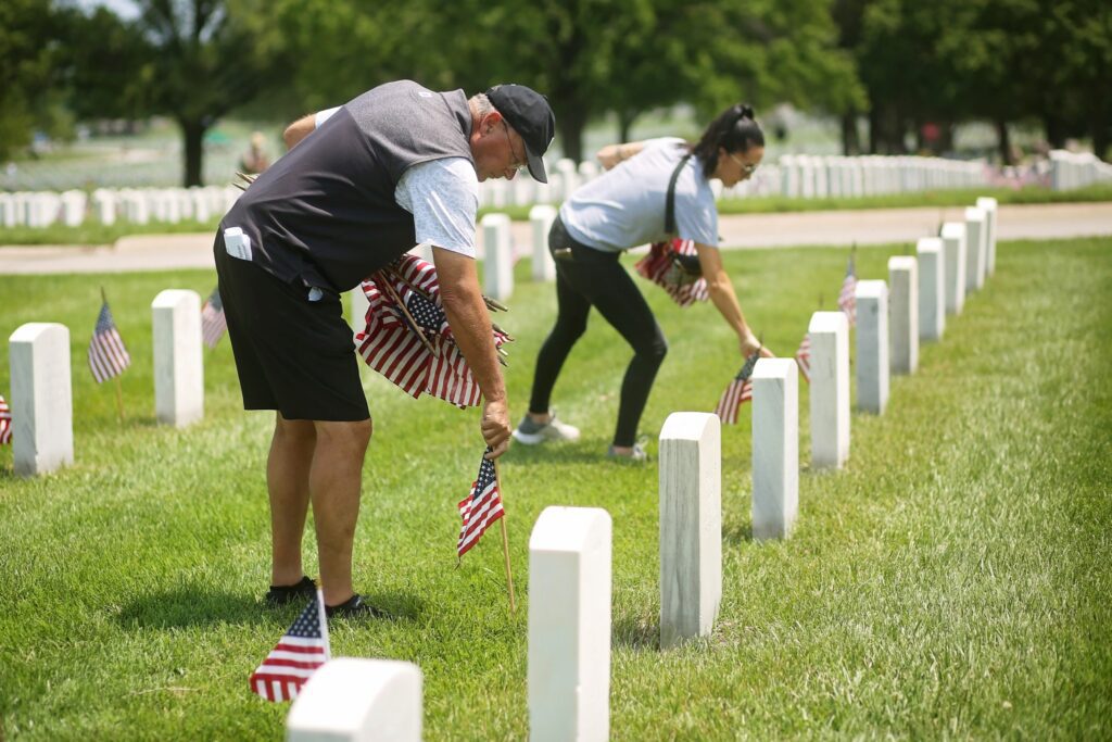 Two people placing small American flags in front of white gravestones in a cemetery on a sunny day. The man in the foreground is wearing a black cap, black shorts, and a gray vest over a white shirt, while the woman in the background is dressed in black pants and a light gray shirt. The gravestones are arranged in neat rows on green grass.