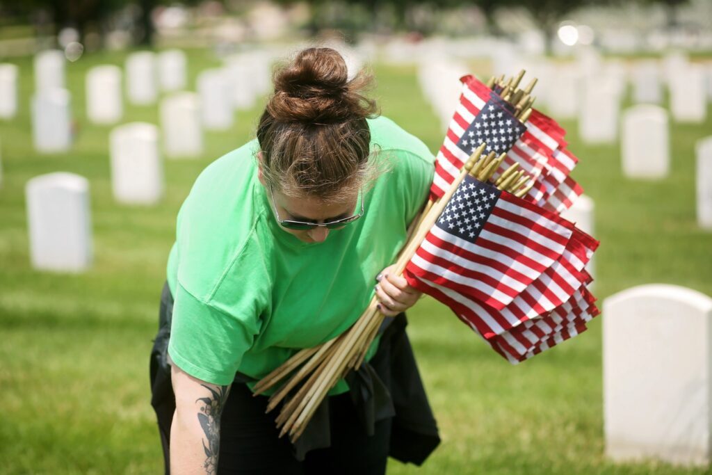 A person wearing a bright green shirt and sunglasses is placing small American flags on wooden sticks in a cemetery with rows of white headstones. The person has their hair tied up in a bun and a visible tattoo on their forearm.