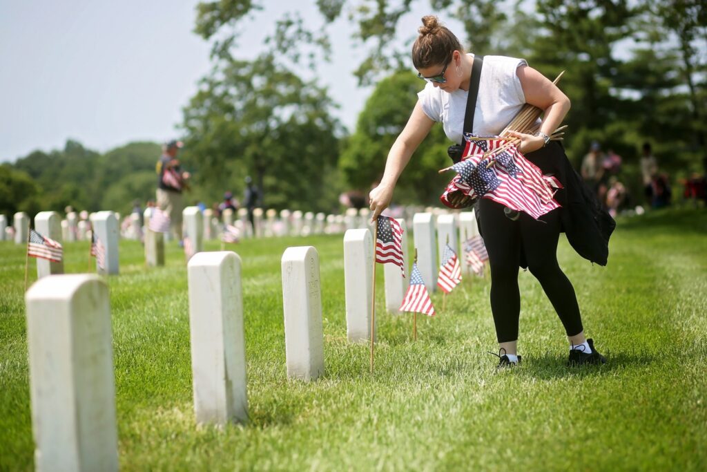 A woman wearing sunglasses, a white sleeveless top, black leggings, and black shoes is placing small American flags next to white gravestones in a cemetery. She holds several flags in her left arm and is bending down to plant one flag in the ground. The cemetery has rows of white gravestones and green grass, with more people in the background also placing flags. Trees and a clear sky are visible in the background.
