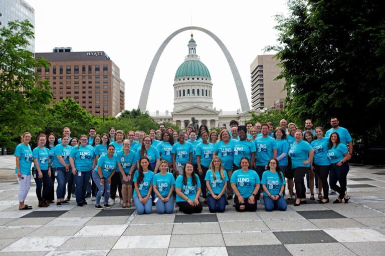 A large group of people wearing matching bright blue "Lung Force" t-shirts posing outdoors on a tiled plaza. Behind them is a historic building with a green dome and the Gateway Arch in the background, indicating the location is St. Louis. The group is smiling and standing closely together, with some kneeling in the front row. Trees and city buildings frame the scene.