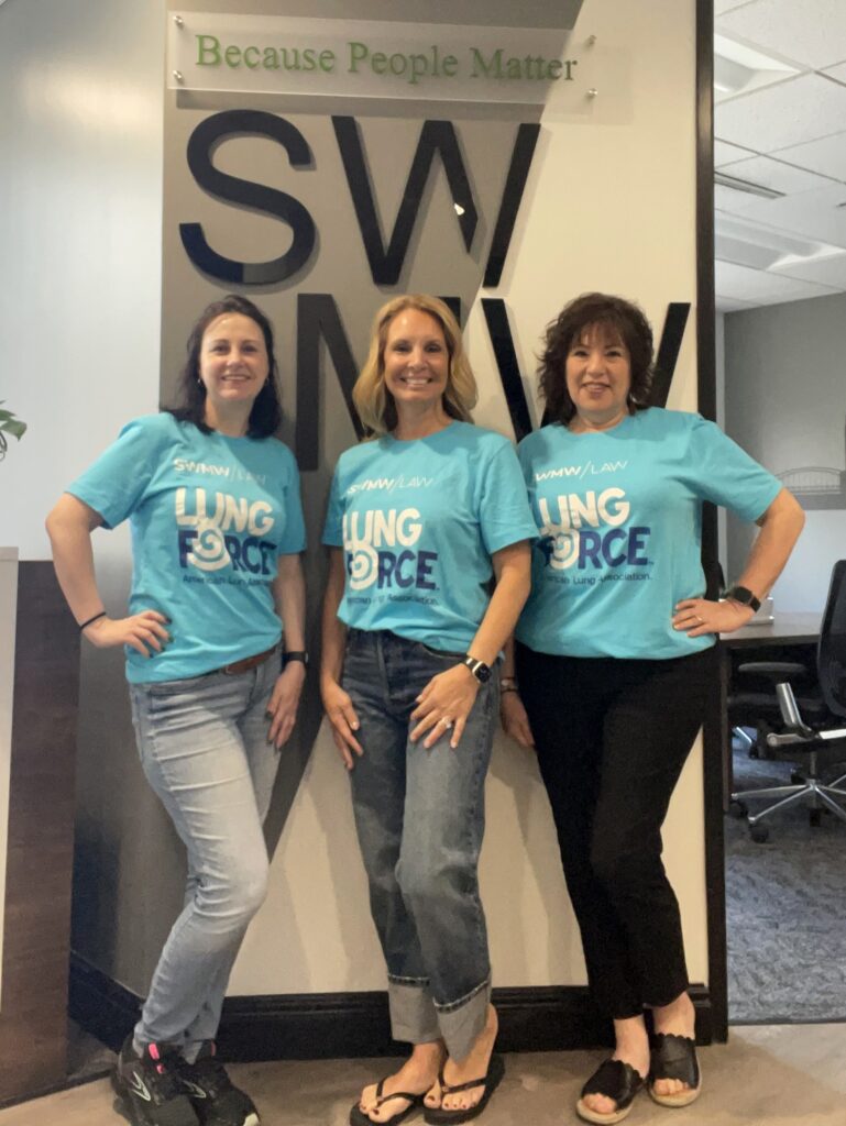 Three women stand side by side in front of a wall with large black letters and a sign above that reads "Because People Matter." They are all wearing matching light blue T-shirts with the text "SWMW LAW LUNG FORCE American Lung Association." The woman on the left is wearing light blue jeans and black sneakers, the woman in the middle is wearing rolled-up blue jeans and black flip-flops, and the woman on the right is wearing black pants and black slip-on shoes. All three have their hands on their hips and are smiling.