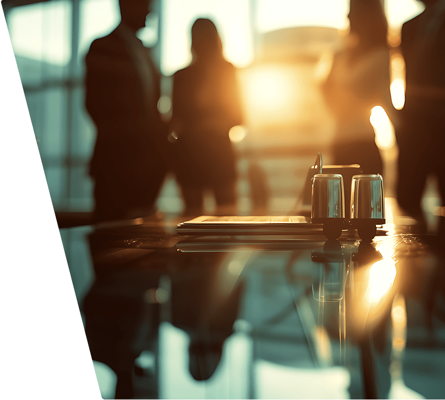 A glossy conference table with a stack of folders and two upside-down glasses on a holder, illuminated by warm sunlight. In the background, four blurred figures stand near large windows, creating a professional office atmosphere.