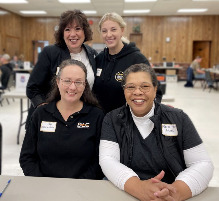 Four women smiling and posing together indoors at a table. Two women are seated in front, one wearing glasses and a black polo shirt with a name tag that reads "Lisa Bloomfield," and the other wearing glasses, a black vest over a white long-sleeve shirt, with a name tag that reads "Roxanne Morris." Behind them stand two women, one with short dark hair wearing a black jacket, and the other with blonde hair in a ponytail wearing a black hoodie with a logo and a name tag that reads "Paige." The background shows a room with wood-paneled walls and other people seated at tables.