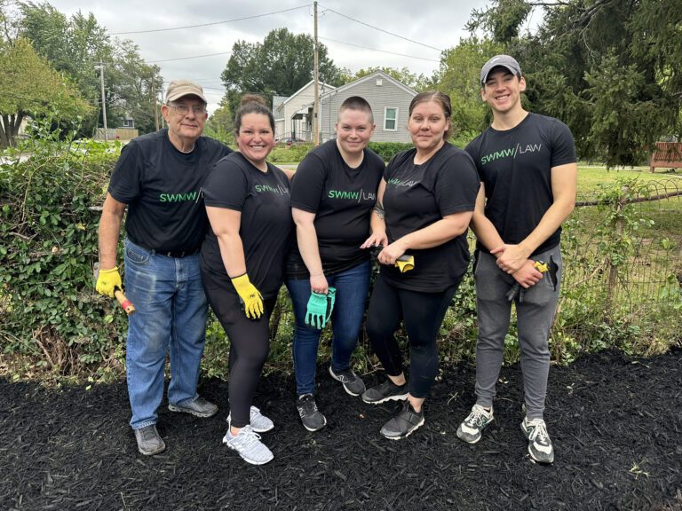 Five people stand outdoors on black mulch in front of a wire fence with greenery. They are wearing matching black t-shirts with "SWMW | LAW" printed on them, along with gloves and casual pants. The group appears to be posing for a photo during a community or volunteer event.