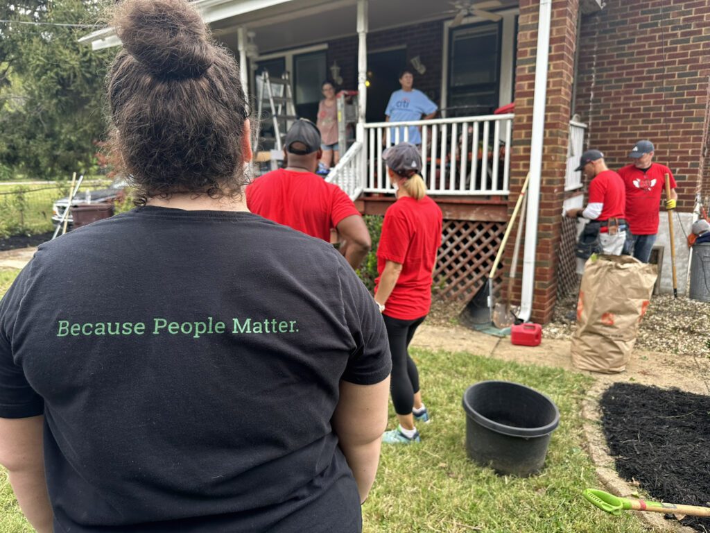 A group of people gathered outside a brick house with a porch. In the foreground, a person with curly hair tied in a bun is wearing a black shirt with the text "Because People Matter." Several others in red shirts are standing and working near the porch, with gardening tools, a large paper yard waste bag, and a black plastic bucket visible on the grass. Two people are standing on the porch talking to the group. The scene suggests a community or volunteer activity.