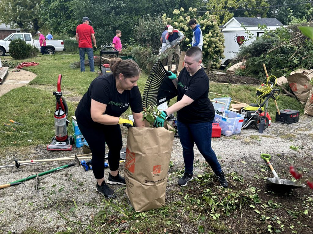 Two people wearing black shirts and gloves are placing garden debris into a large brown paper yard waste bag. Various gardening tools, equipment, and supplies are scattered around them on a gravel and grassy area. In the background, several other people are engaged in yard work near bushes and trees, with a white truck and a trailer visible.