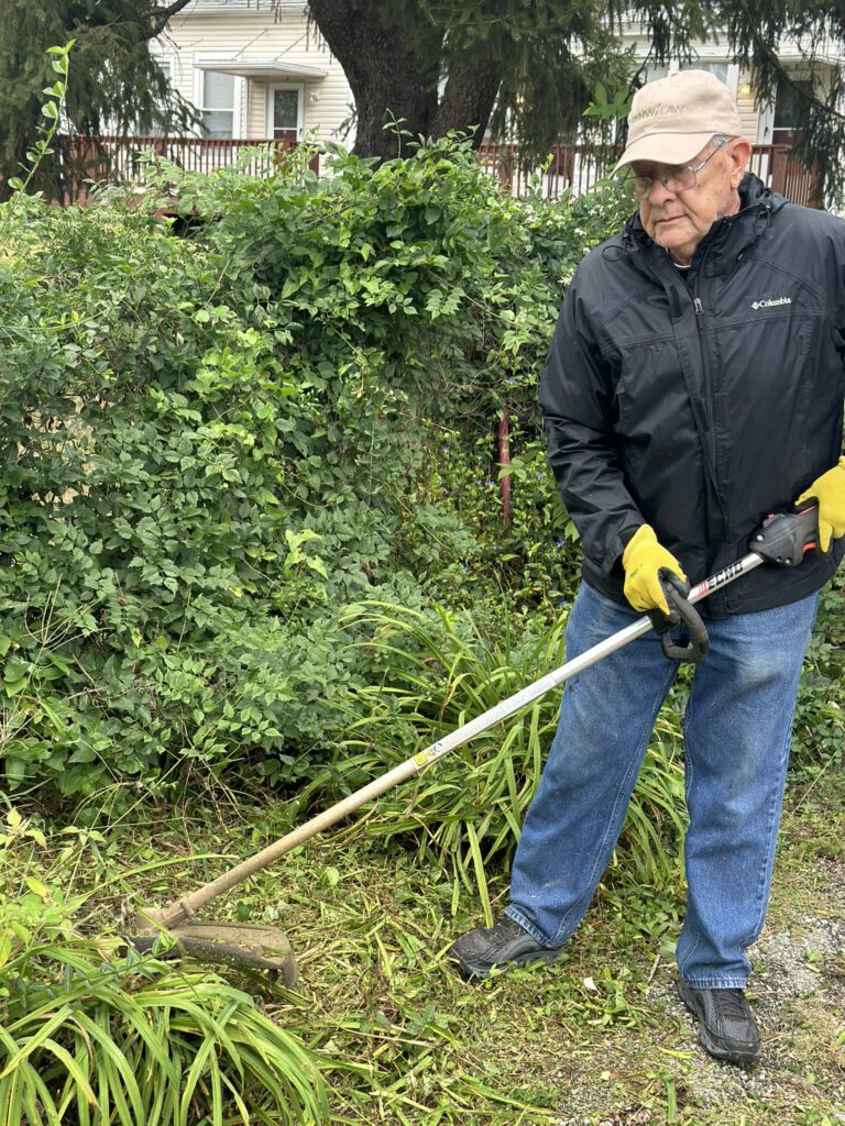 An elderly man wearing a beige cap, glasses, a black Columbia jacket, yellow gloves, blue jeans, and black shoes is using a string trimmer to cut grass and weeds near a dense green bush in a garden. A house with a porch and windows is visible in the background.