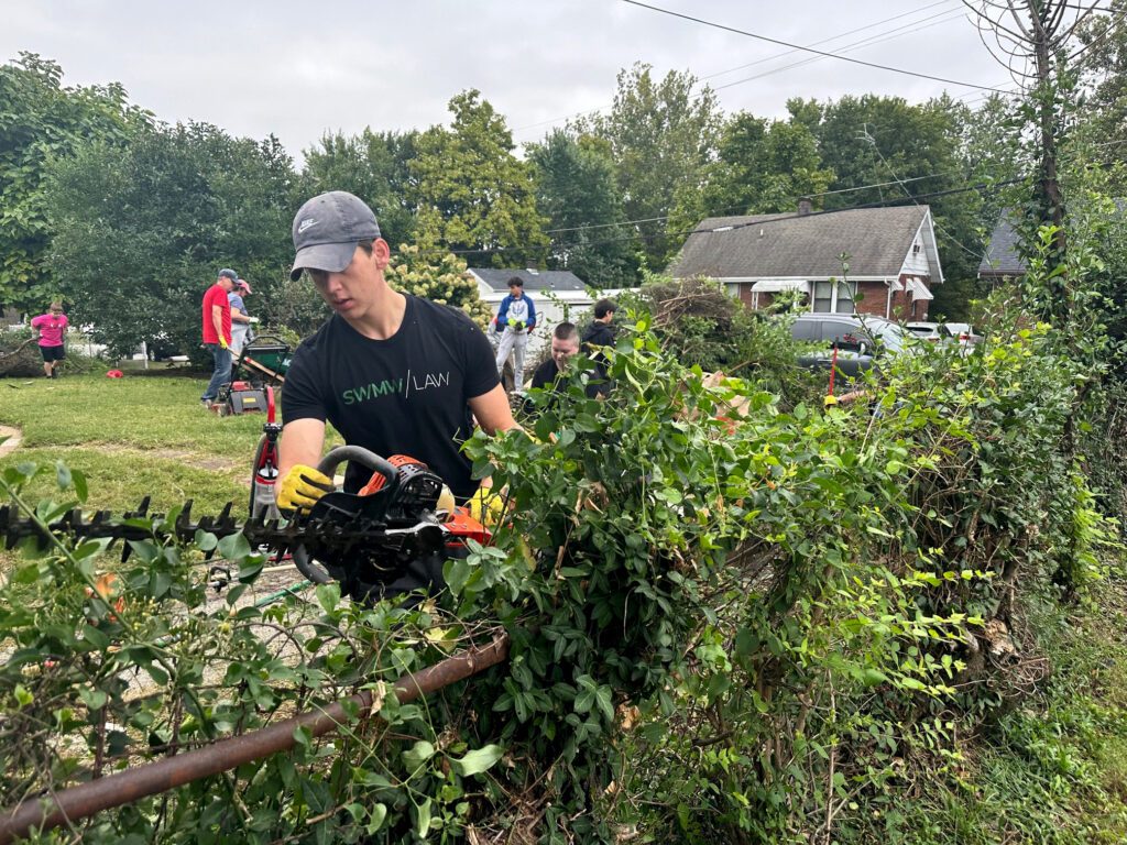 A young man wearing a gray Nike cap, black "SWMM LAW" t-shirt, and yellow gloves is trimming overgrown bushes with a hedge trimmer along a rusty metal fence. In the background, several people are engaged in yard work, with green trees, grass, and houses visible under a cloudy sky.