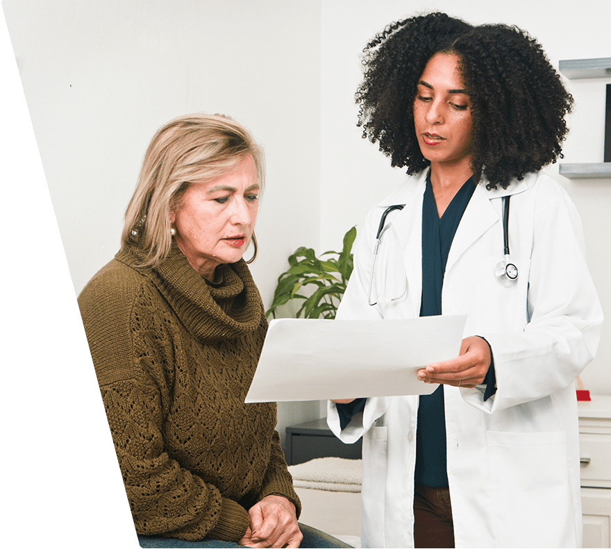A female doctor with curly hair wearing a white coat and stethoscope is showing a document to an older woman in a brown knitted sweater, who is sitting and attentively looking at the paper. They are in a clinical setting with a plant and shelves in the background.