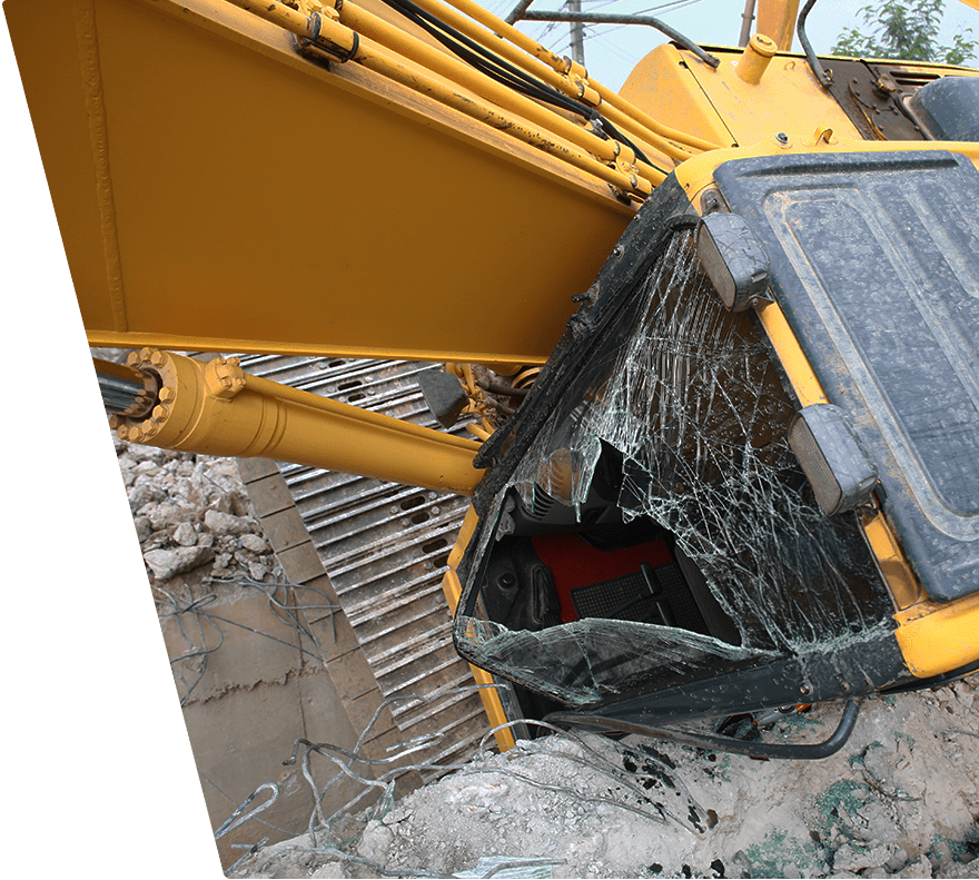 Yellow excavator with a severely shattered front windshield, showing cracks and a large hole. The excavator is positioned on rocky and concrete debris, with visible hydraulic arms and metal tracks.
