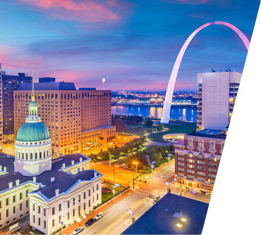 A cityscape at dusk featuring the illuminated Old Courthouse with a green dome in the foreground, modern buildings, and the iconic Gateway Arch in the background near a river. The sky is a gradient of blue and pink hues, and streetlights and building lights are glowing warmly.