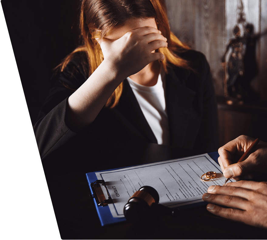 A woman with light brown hair, wearing a black blazer and white shirt, is sitting at a table with her hand covering her face. On the table, there is a clipboard with a contract, two gold wedding rings, a gavel, and a person's hands holding a pen, appearing to sign the document. The background includes a wooden wall and a statue.