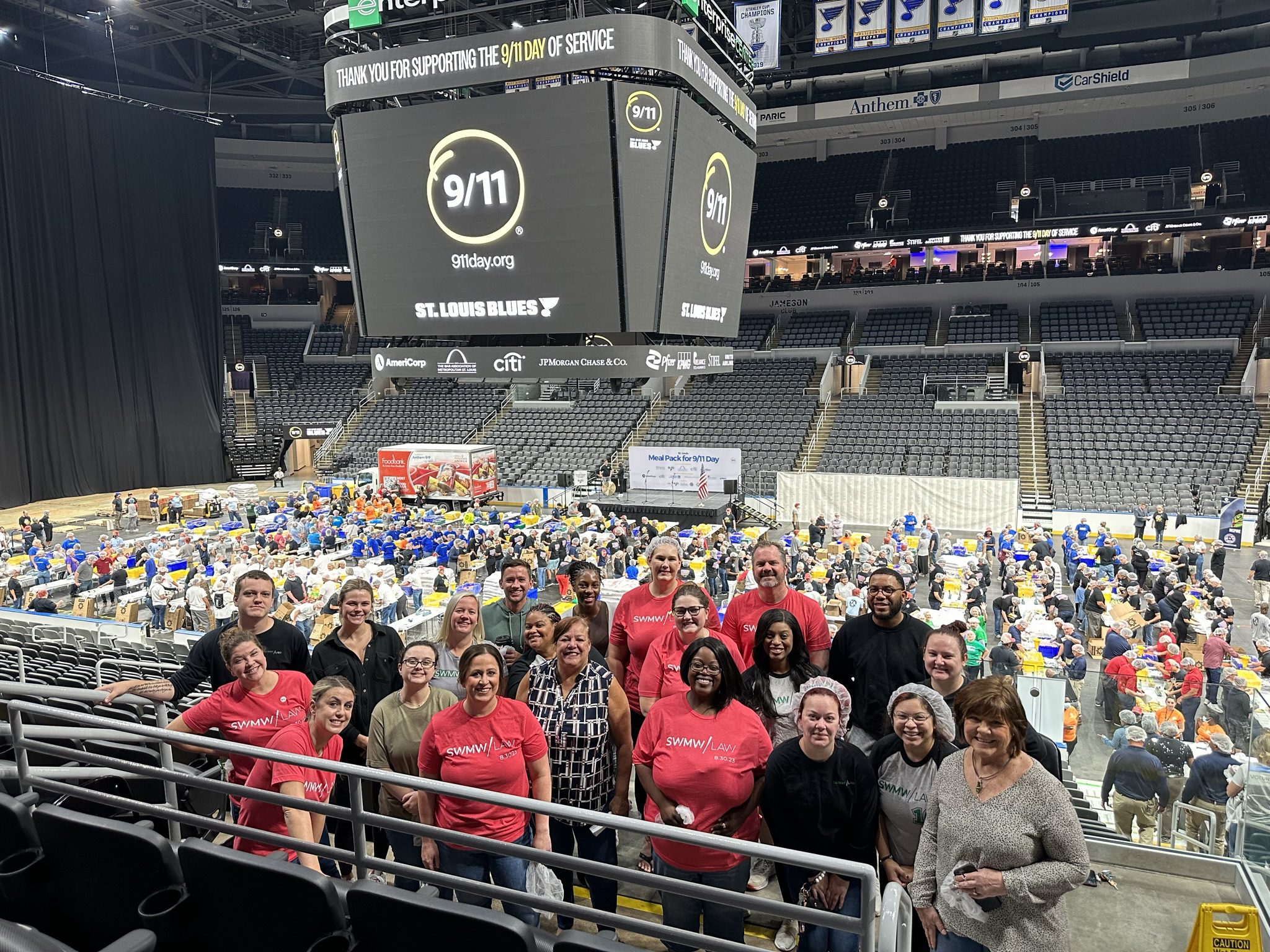 A group of people posing for a photo inside an arena with many volunteers working at tables in the background. The arena scoreboard displays a message supporting the 9/11 Day of Service with the text "9/11 911day.org ST. LOUIS BLUES." Several individuals in the group wear red and gray shirts with "SWMW | LAW" printed on them. The arena seats are mostly empty, and a banner in the background reads "Meal Pack for 9/11 Day.