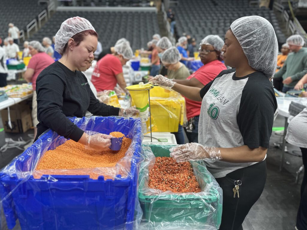 Two women wearing hairnets and gloves are packing food items in a large indoor space. One woman is scooping red lentils from a large blue bin, while the other is holding a yellow funnel over a green bin filled with dried vegetables. Several other people in the background are also engaged in similar packing activities.