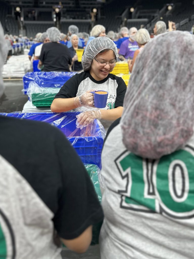 A group of people wearing hairnets and gloves are working together in an assembly line, packaging food. One person in the center is smiling and holding a blue cup filled with food, standing behind a blue plastic bin lined with a plastic bag. Many others in the background are similarly engaged in the packing process.