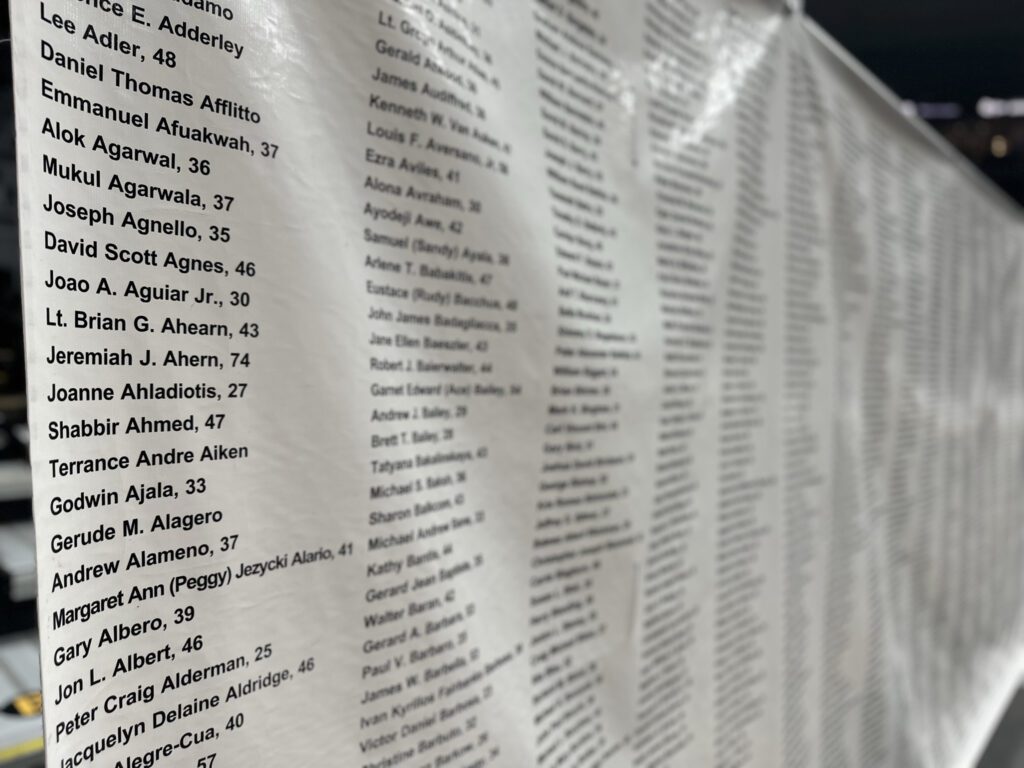 A close-up of a memorial wall with numerous names listed in black text on a light-colored surface. The names are organized in columns, with some including ages next to them. The focus is sharp on the left side, showing names such as Lee Adler, Daniel Thomas Afflitto, Alok Agarwal, and others, while the right side gradually blurs into the background. The setting appears to be outdoors, possibly at a commemorative site.