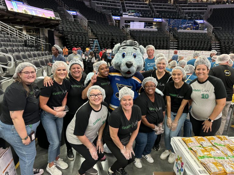 A group of people wearing hairnets and black or gray "SWMW | LAW" t-shirts pose together indoors on a sports arena floor. They are smiling and some wear plastic gloves. In the center is a person in a blue and yellow St. Louis Blues mascot costume. A table with packaged food items is visible in the foreground on the right. Empty stadium seats and other people are in the background.