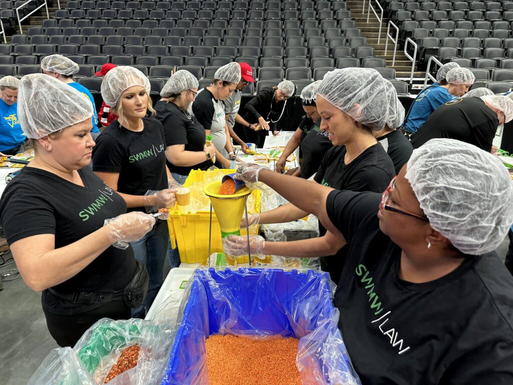 A group of people wearing black "Swimmy Law" t-shirts, hairnets, and plastic gloves are working together to measure and pour orange lentils into a yellow funnel over a blue bin. They are standing at tables with large bins of lentils and other supplies, in what appears to be an indoor arena with empty gray seats in the background.