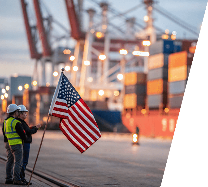 An American flag on a pole held by a person wearing a yellow safety vest and white hard hat, standing next to another person in a white hard hat at a shipping port with stacked cargo containers and cranes in the background.