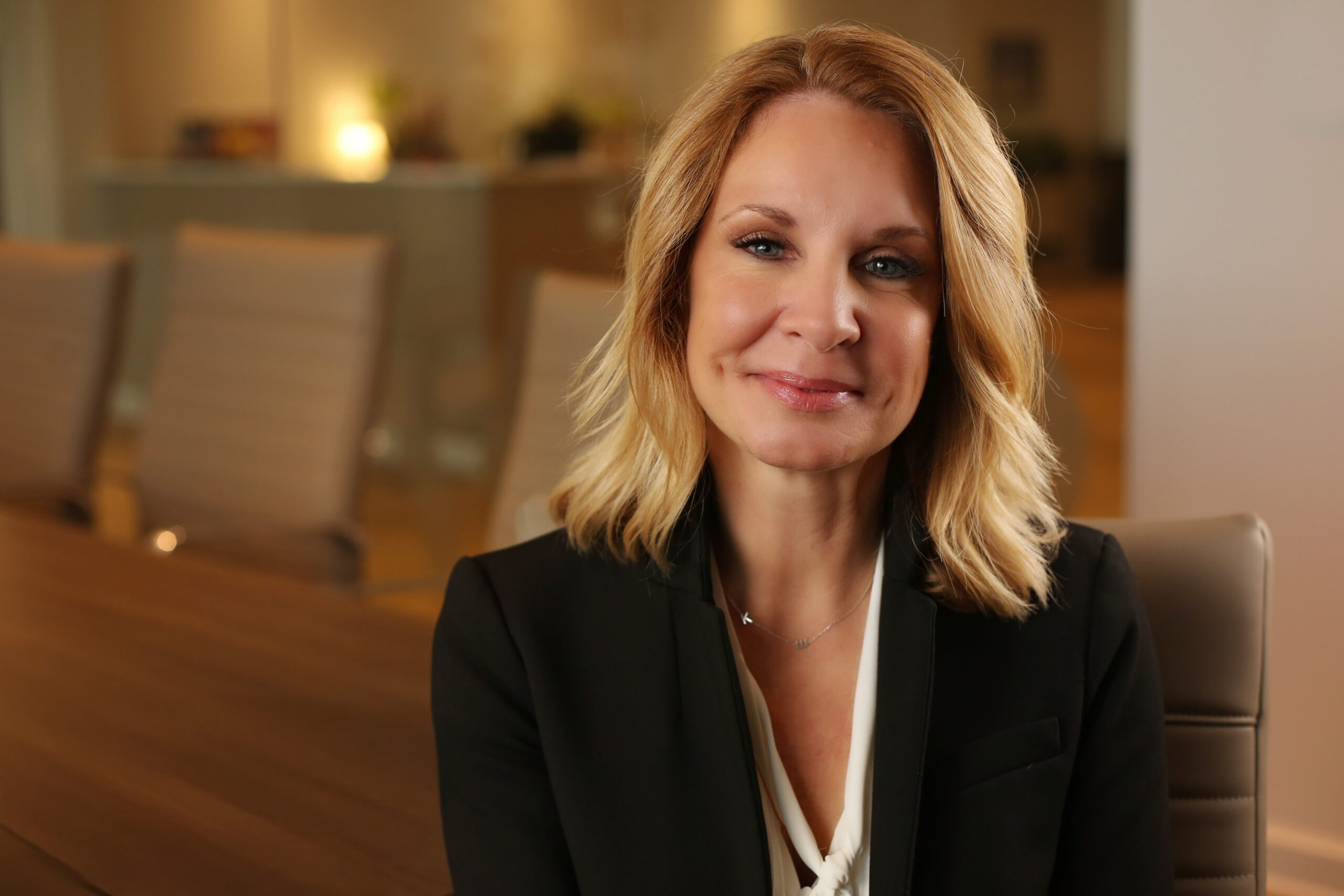 Blonde woman with shoulder-length hair wearing a black blazer and white blouse, sitting in a modern office setting with beige chairs and a wooden table in the background. She has a subtle smile and is wearing a delicate necklace. The background is softly lit and out of focus.