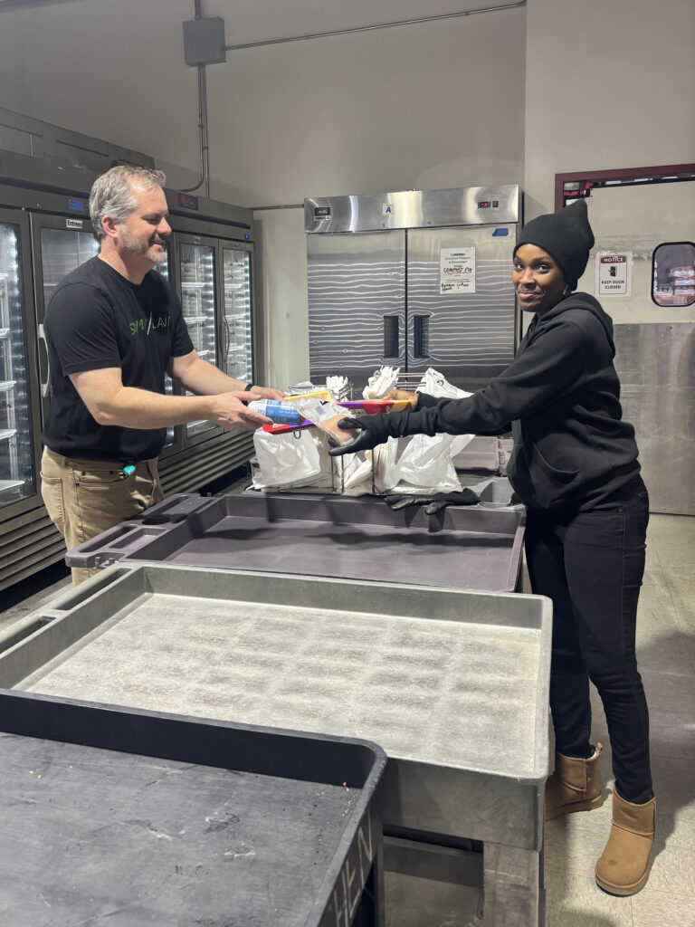 A man and a woman are exchanging packaged food items in a commercial kitchen or food storage area. The man, wearing a black t-shirt and khaki pants, is handing food to the woman, who is dressed in a black hoodie, black pants, a black beanie, black gloves, and tan boots. Behind them are large stainless steel refrigerators and freezers, with plastic bags and trays on the counter between them.
