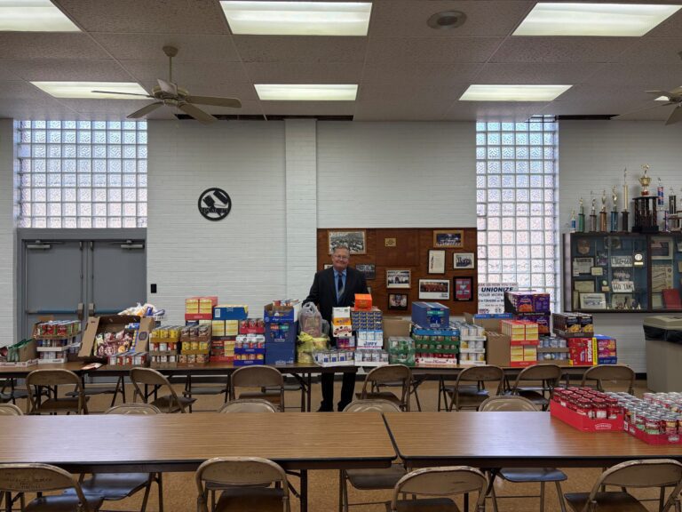 A man in a suit stands behind a long table filled with various canned and boxed food items, including pasta, canned vegetables, and snacks. The room has white brick walls, glass block windows, ceiling fans, and fluorescent lights. There are folding chairs around the tables, and a trophy case and bulletin board are visible on the right wall.