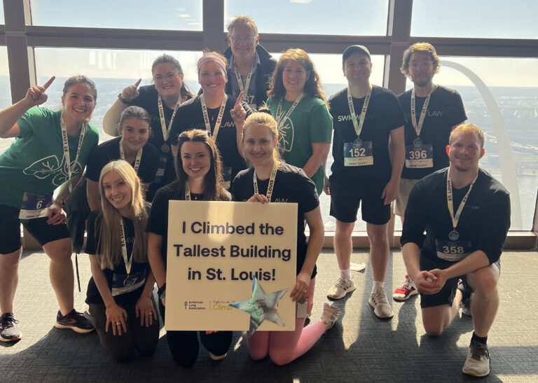 A group of twelve people posing indoors in front of large windows with a view of the St. Louis Gateway Arch. Most are wearing black shirts with "LAW" or "SWIM" printed on them, and some wear green shirts with a lung graphic. Many have medals around their necks and race bibs on their shorts. Two women kneeling in front hold a sign that reads, "I Climbed the Tallest Building in St. Louis!" The group is smiling and some are holding up one finger in a celebratory gesture.