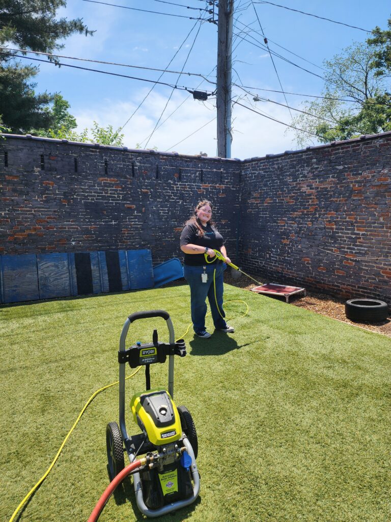 A person standing on artificial grass in a fenced outdoor area, holding a yellow pressure washer wand connected to a Ryobi pressure washer machine in the foreground. The background features a brick wall, some wooden boards leaning against it, a tire, and a small raised platform. The sky is clear with some power lines visible above.
