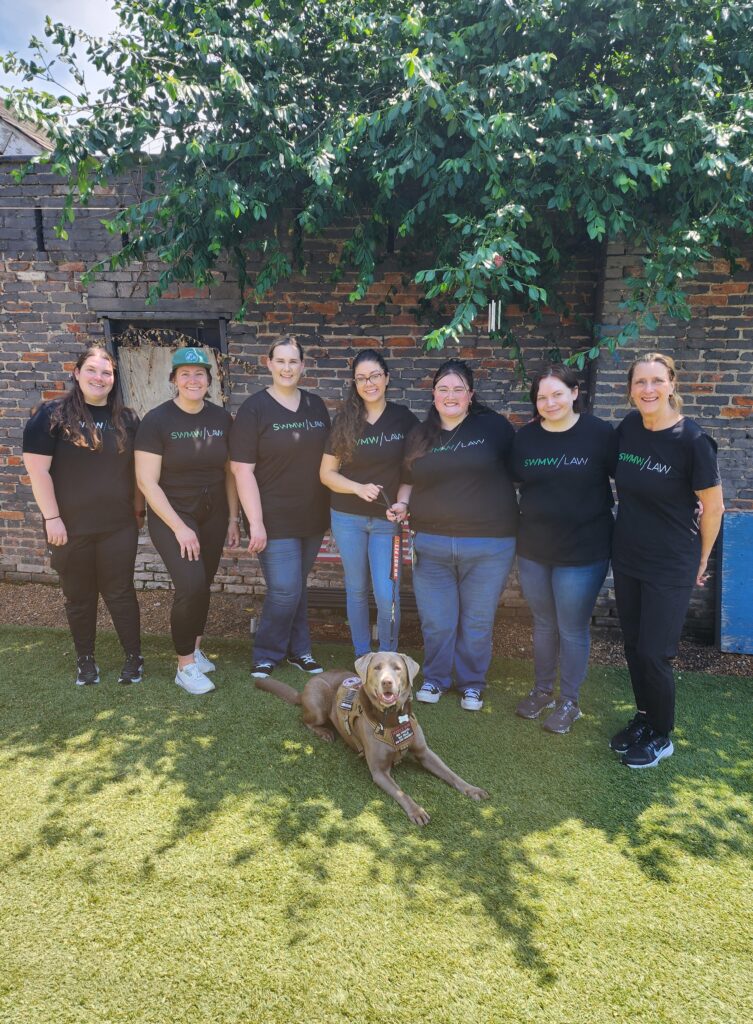 Seven women wearing black "SWMW | LAW" t-shirts stand in a row outdoors on grass in front of a brick wall with greenery above. A brown service dog with a harness lies on the grass in front of them.