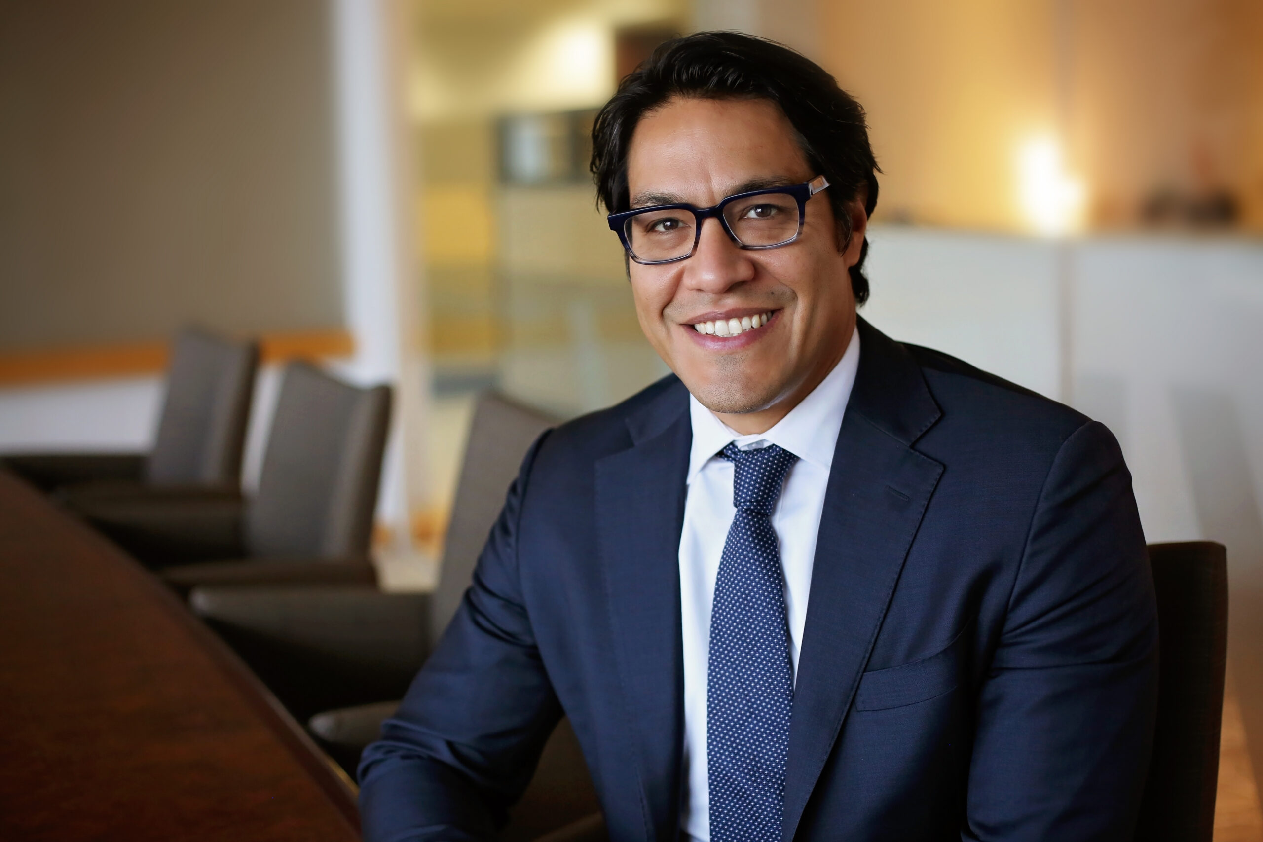 Man with dark hair and glasses wearing a navy blue suit, white shirt, and patterned tie, smiling while seated at a conference table in a modern office setting.