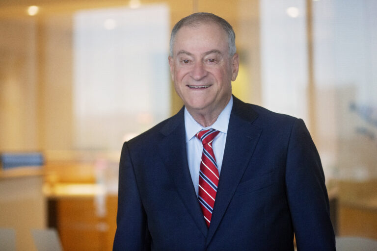 Middle-aged man wearing a dark navy suit, light blue dress shirt, and a red striped tie, smiling in an office setting with blurred background.
