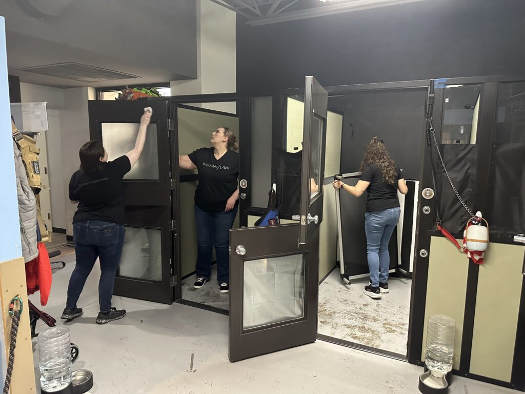 Three women are cleaning and organizing a room with black-framed glass doors. One woman on the left is wiping a door, the woman in the middle is cleaning the inside of a small room, and the woman on the right is moving a folding table. The room has a light gray floor and light-colored walls with black trim. Various items, including coats and a water dispenser, are visible around the space.